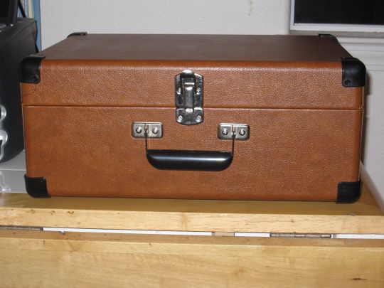 Photo shows the Crosley CR249 turntable on a table. The cover is closed. A carrying handle is visible.