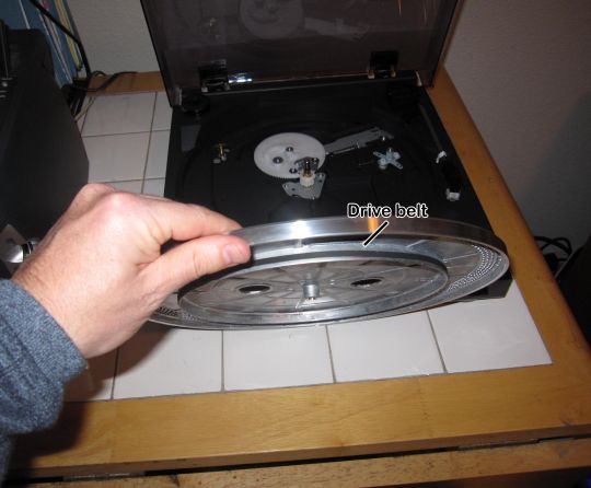 Photo shows a hand holding up a platter with a rubber drive belt wrapped around the bottom. The unassembled Audio-Technica AT-LP2D-USB turntable is in the background.